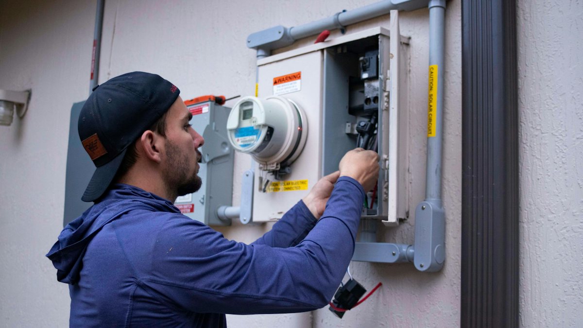 Electrician in a cap and hoodie working on an electrical meter outside a building